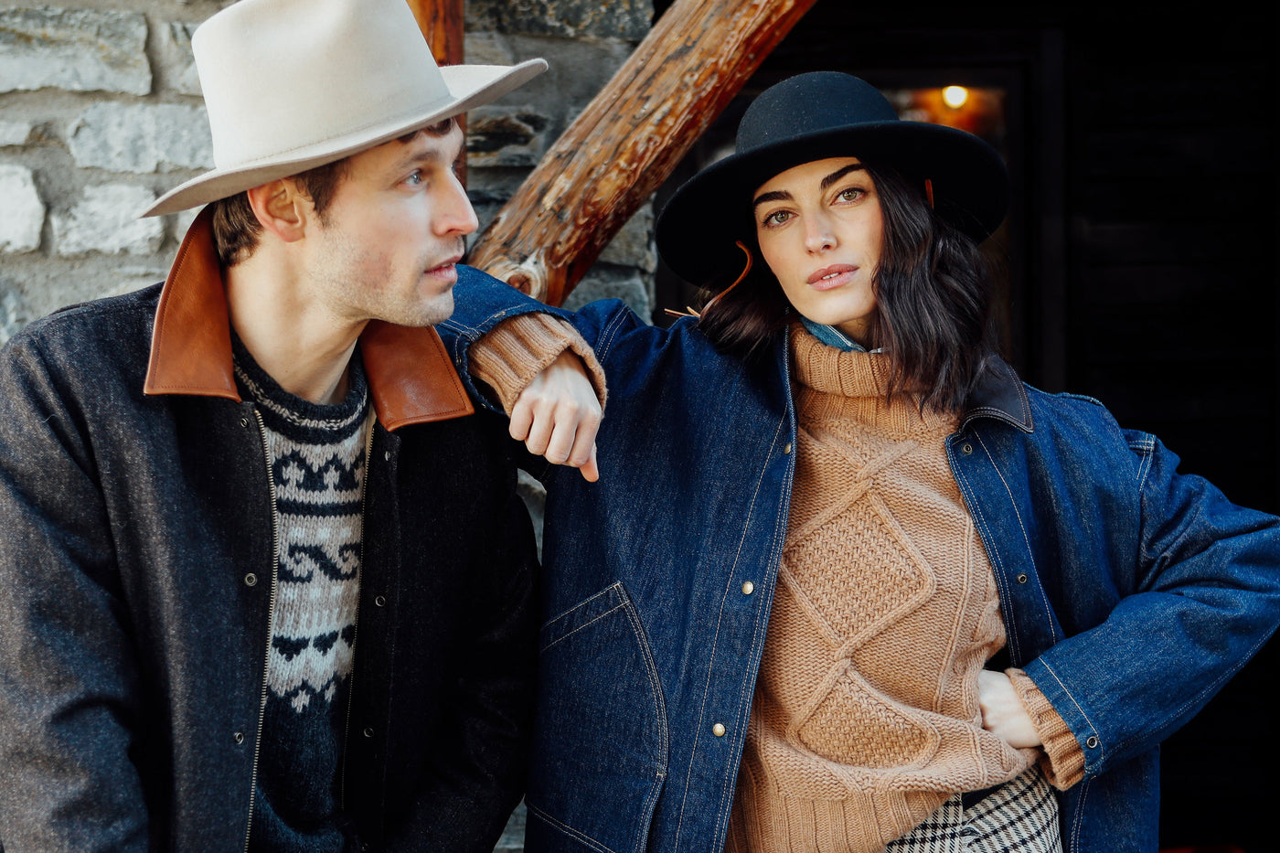 Two people standing against a stone wall, one wearing a hat and coat, the other in a sweater and plaid skirt.